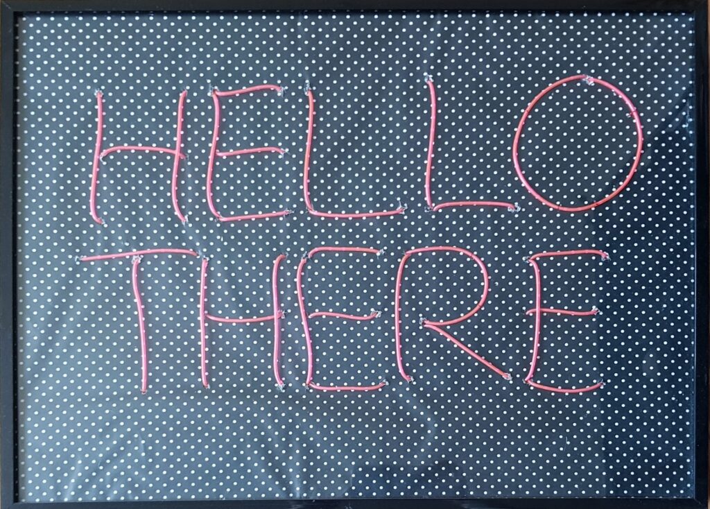 Ein Foto von einem Schild mit dem Schriftzug "HELLO THERE" in rosa Buchstaben auf schwarzem Hintergrund mit kleinen weißen Punkten in einem schwarzen Rahmen.