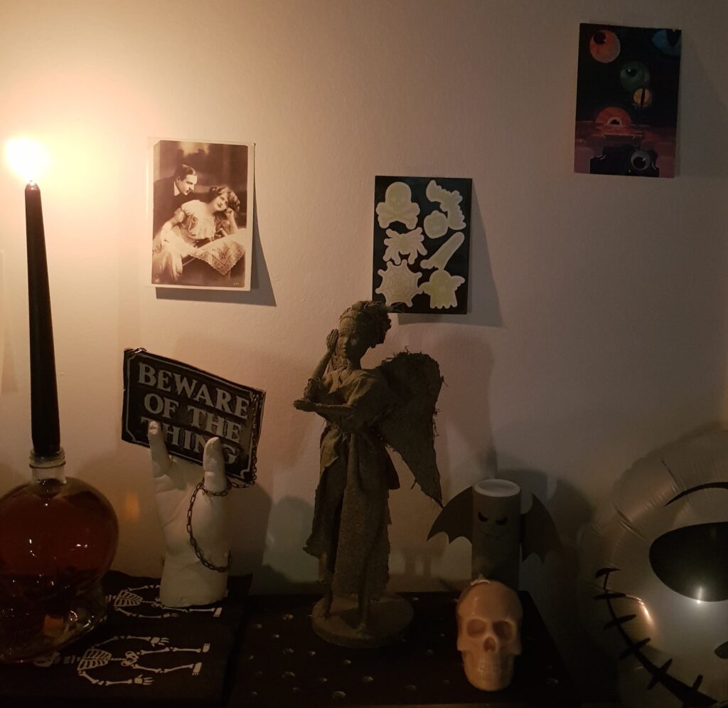 Verschiedene Halloweendekoelemente stehen zusammen. Darunter ein gläserner Totenkopf mit schwarzer Kerze, eine Gipshand mit einem Schild an einer Kette auf dem "Beware of the Thing" steht und eine "Weeping Angel" Figur.