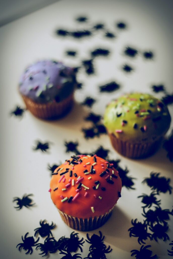 Auf einem weißen Untergrund befinden sich drei bunt überzogene Muffins mit bunte Zuckerstreuseln in schwarz, grün und pink. Der Muffin ganz vorne ist knallorange. Im Hintergrund befinden sich ein giftgrüner und violetter Muffin. Rund herum liegen kleine Plastikspinnen.