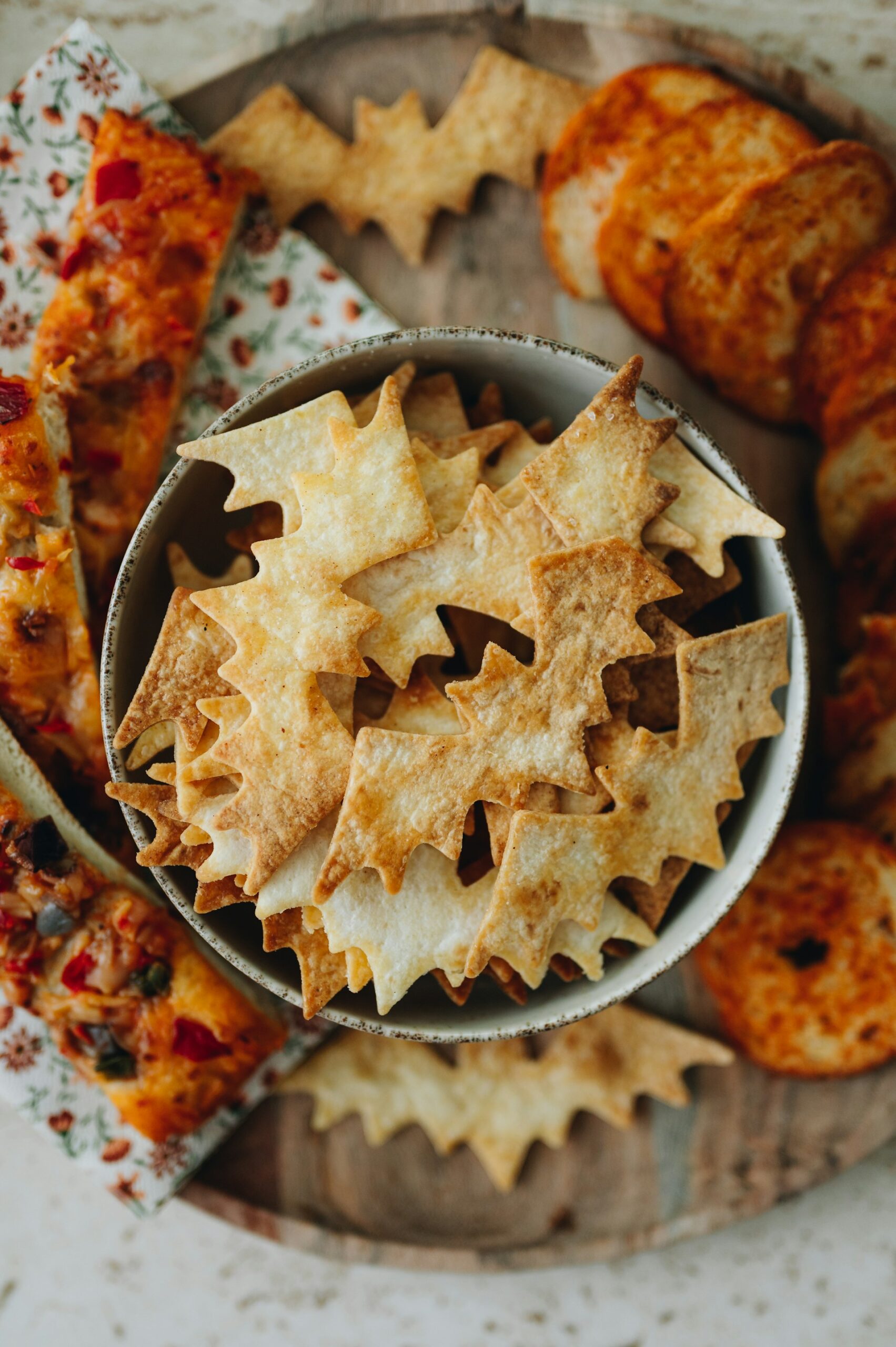 Ein Schüsselchen mit Fledermausgebäck auf hölzernem Tablett mit geblümten Servietten und herzhaften anderen Snacks rund herum.