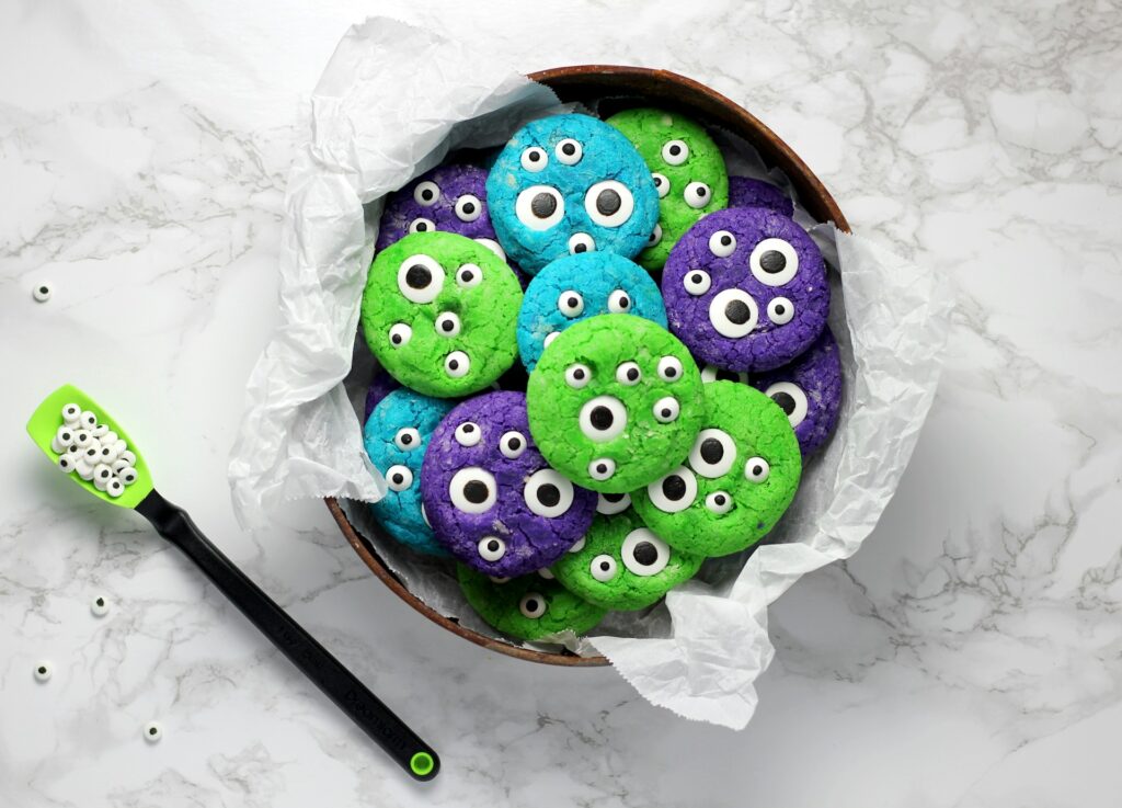 Eine Schüssel mit giftgrün, leuchttürkis und neonviolett eingefärbten runden Keksen mit Zuckeraugen steht auf einer Marmoroberfläche, aus der Schüssel ragt etwas helles Backpapier, daneben liegt ein giftgrüner Löffel mit Zuckeraugen.