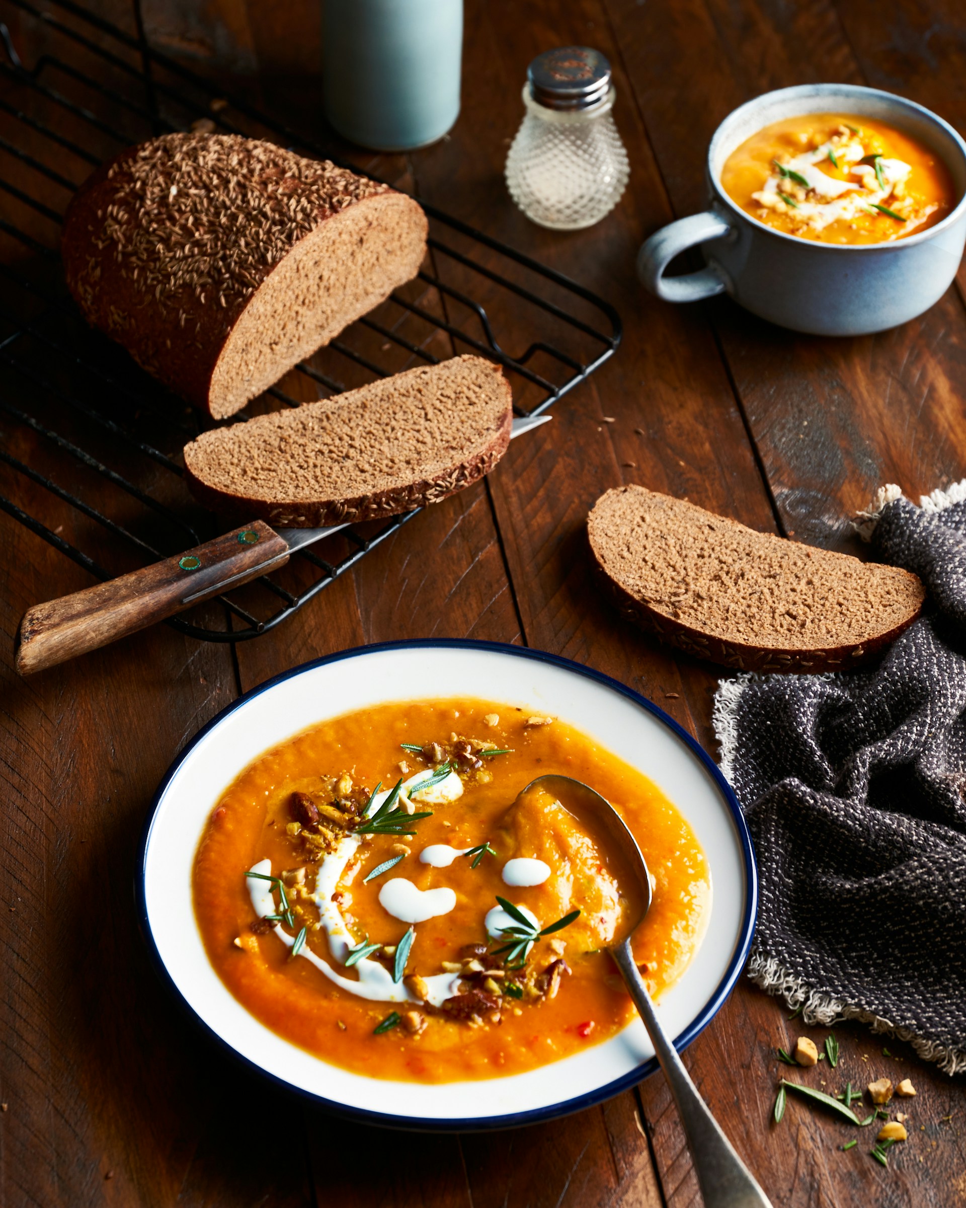 Auf einem Holztisch steht ein Teller Kürbissuppe mit Löffel, hübsch garniert mit Rosmarin, Kernen und Sahne. Auf dem Tisch befindet sich noch ein Gitter mit einem halben Laib Brot und zwei abgeschnittenen Scheiben. Auch ein Messer, eine weitere Tasse mit Suppe, ein Salzstreuer und einen Küchentuch sind kunstvoll im Hintergrund auifgebaut.