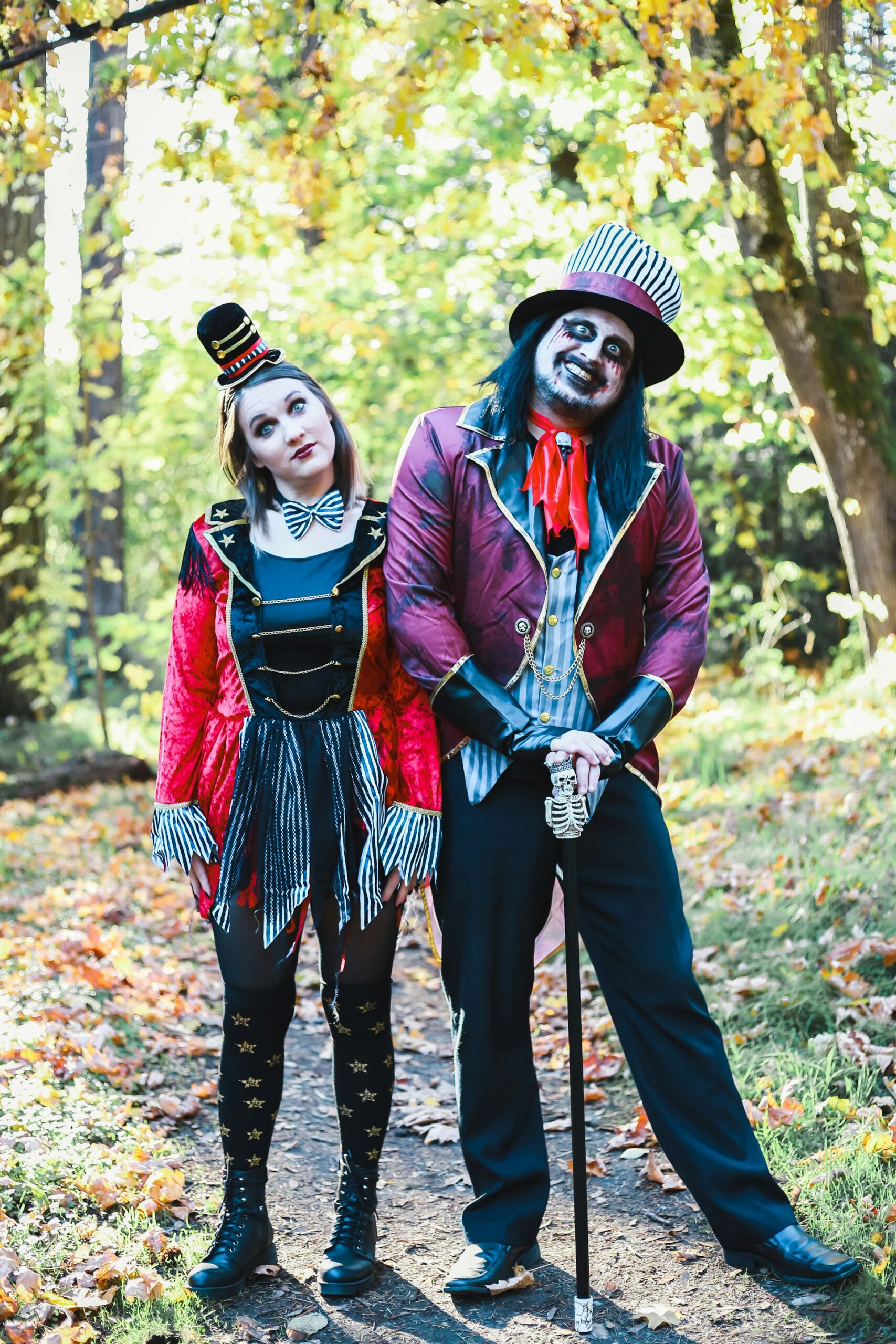 Zwei Menschen in dompteurähnlichen Zirkusartistenoutfits stehen im Wald. Die Frau trägt ein rot-schwarzes Kleid mit schwarzweiß-gestreiften Ärmelaufsätzen und Stoffstreifen am Rock. auf ihrer schwarzen Strupfhose sind diesselben golden Sterne, wie auf dem Revier. Datz trägt sie schwarze Boots, eine schwarz-weiß-gestreifte Fliege und eine winzige schwarze Melone mit roten und schwarz-weißen Streifen und goldenen Schnüren, wie sie auch am Kleidoberteil zu finden sind. Der Mann trägt schwarze Hose und Schuhe, stütz sich auf einen Stock mit Skelettgriff. Dazu trägt er ein dunkelpinkes Dompteursjackett mit schwarz und goldenem Rand an Revier und Ärmeln. Darunter hat er eine Weste an, die blau-weiß gestreift ist. Auf dem Kopf hat einen großen schwarz-weißen Hut mit dunkelpinkem Band und am Hals ein rotes Band. Er ist wie ein unheimlicher Clown geschminkt.