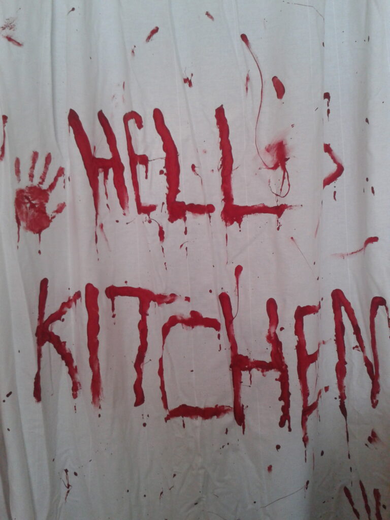Weißer Vorhang auf dem in roten, blutigem Buchstaben "Hell's Kitchen" steht. Dazu ein Handabdruck und Spritzer.