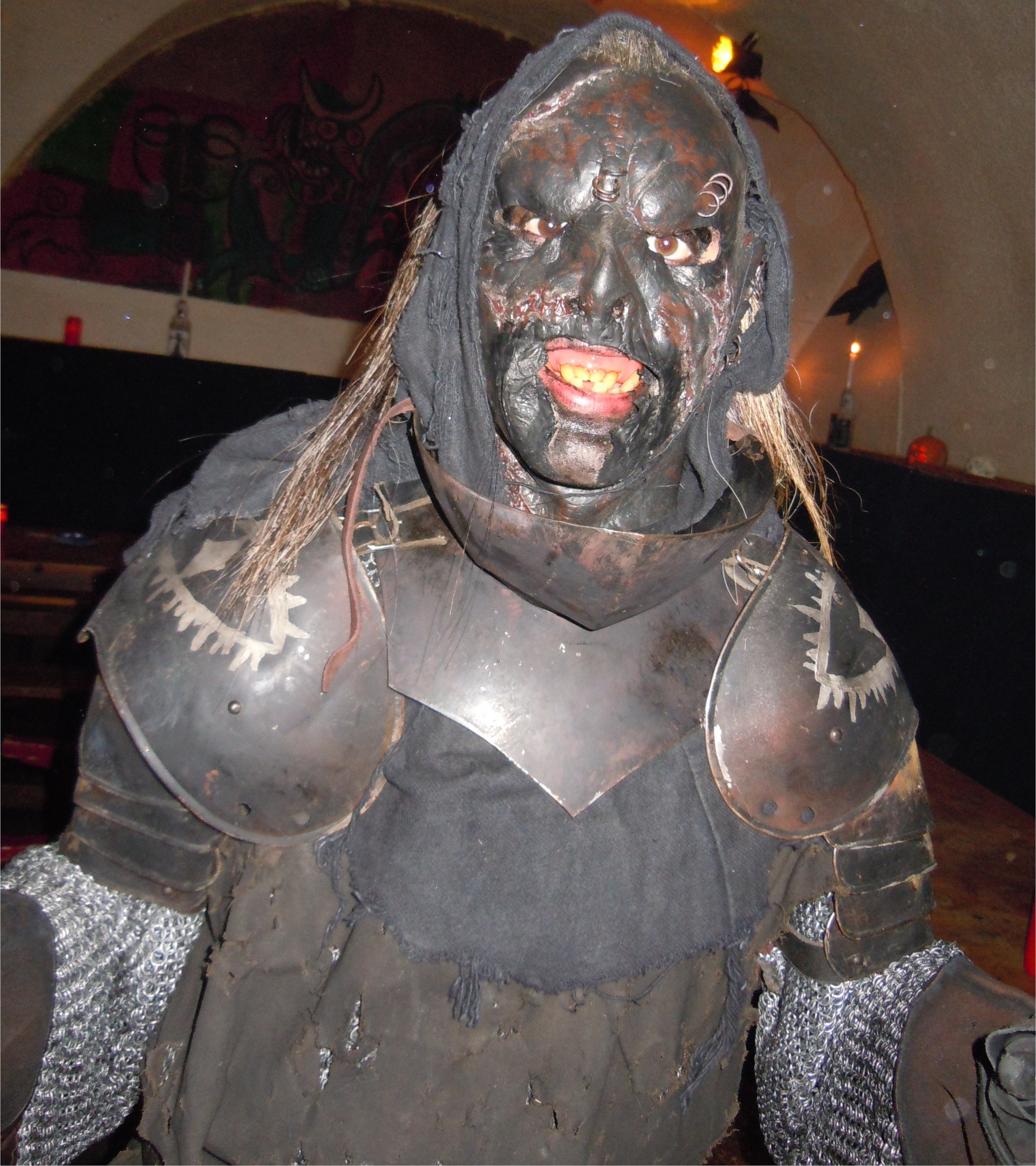 Ein Mensch in einem aufwendigen Live Action Roleplay-Ork-Kostüm: mit Kettenhemd, Schulterrüstung, dunkler Leinenkittel, dunkle Maske mit Furchen, Narben und Piercings, Gebiss mit schiefen gelben Zähnen und zu guter letzt bedeckt ein Tuch mit angeklebten Haarfetzen den Kopf.