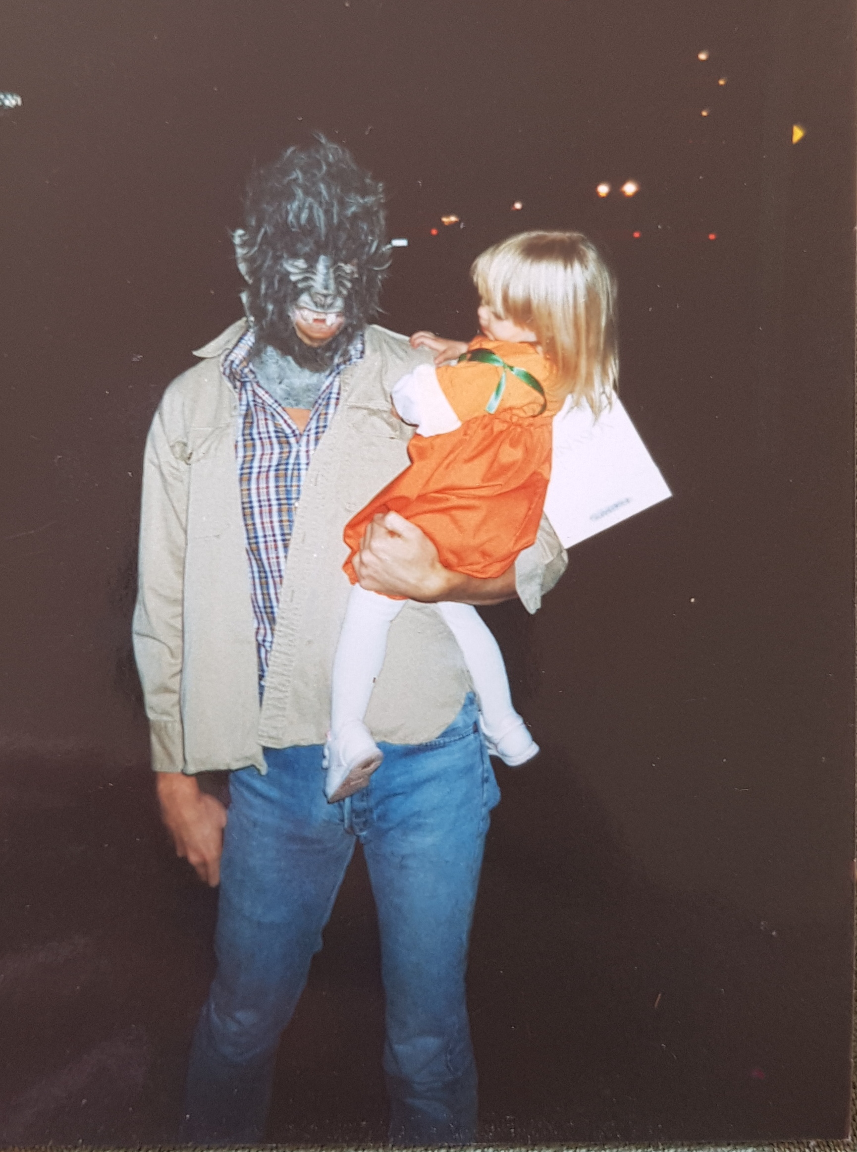 Ein Mann mit einer zotteligen Gorillamaske hat ein blondes Kleinkind, das als Kürbis verkleidet ist auf dem Arm. Er blickt zur Kamera und das Mädchen betrachtet interessiert seine Maske. Sie haben eine weiße Tüte. Der Hintergrund ist dunkel.